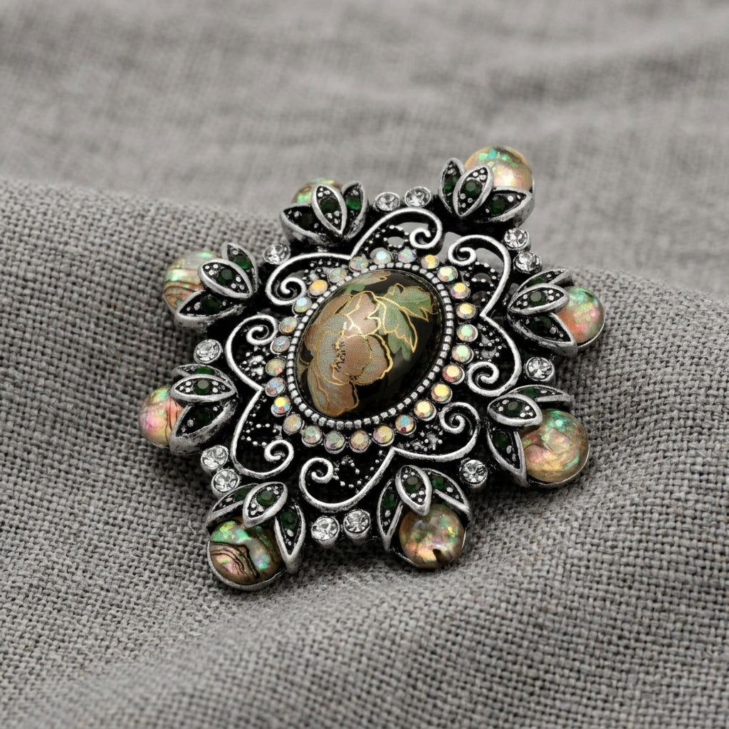 An ornate vintage-style brooch featuring a central oval jet black stone surrounded by filigree, iridescent green and iridescent pink stones, and small AB crystals displayed on a textured gray fabric background.