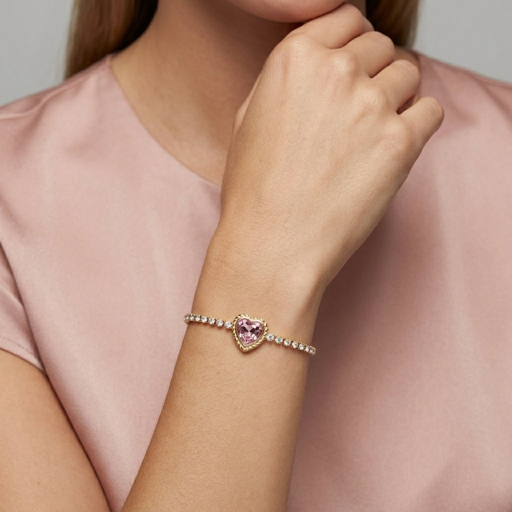 A close-up of a woman wearing a delicate bracelet on her wrist. The bracelet features a central heart-shaped pink gemstone surrounded by small clear stones on a gold-toned band.