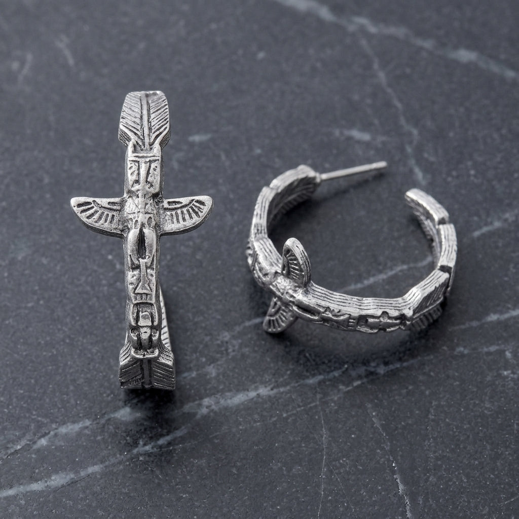 A pair of silver-toned hoop earrings with detailed feather and totem pole motifs, displayed on a dark gray marble surface.