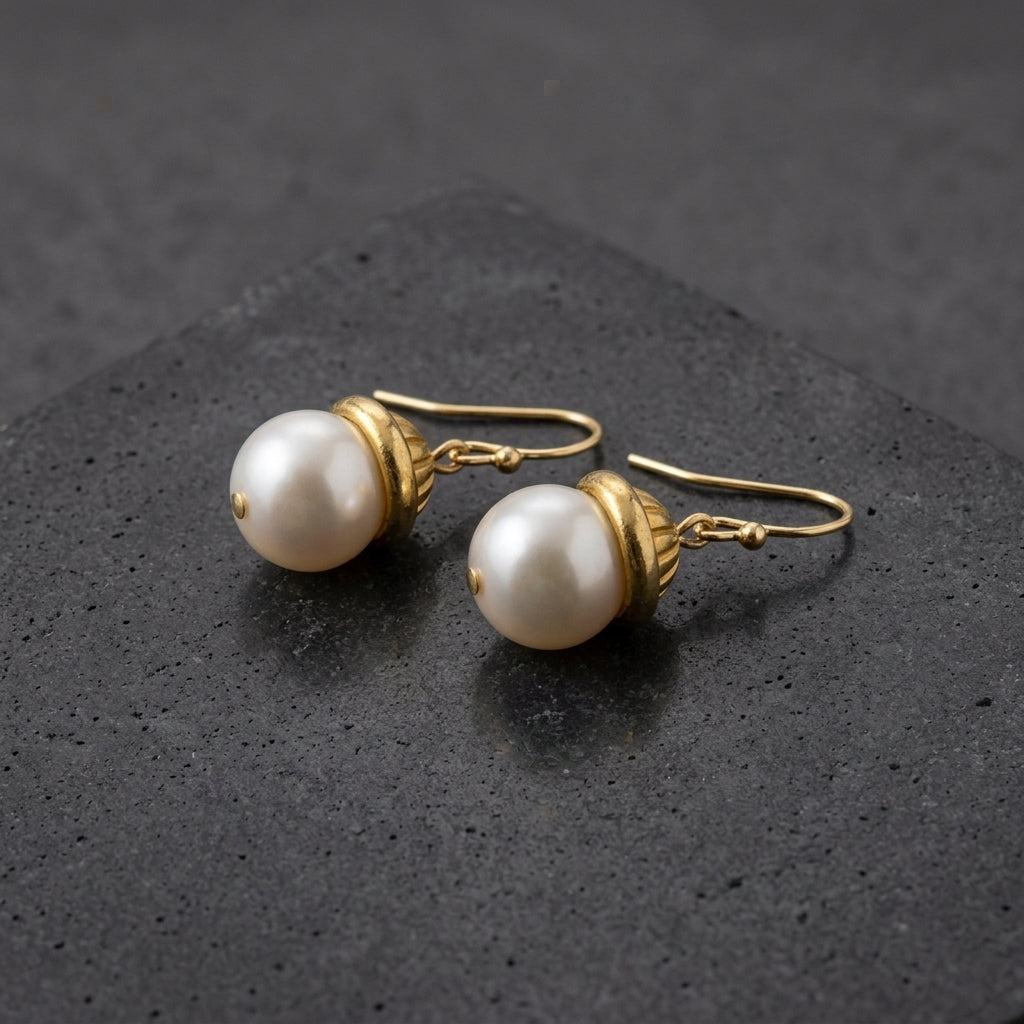 White faux pearl earrings with a gold tone fish hook backing resting on a dark stone surface