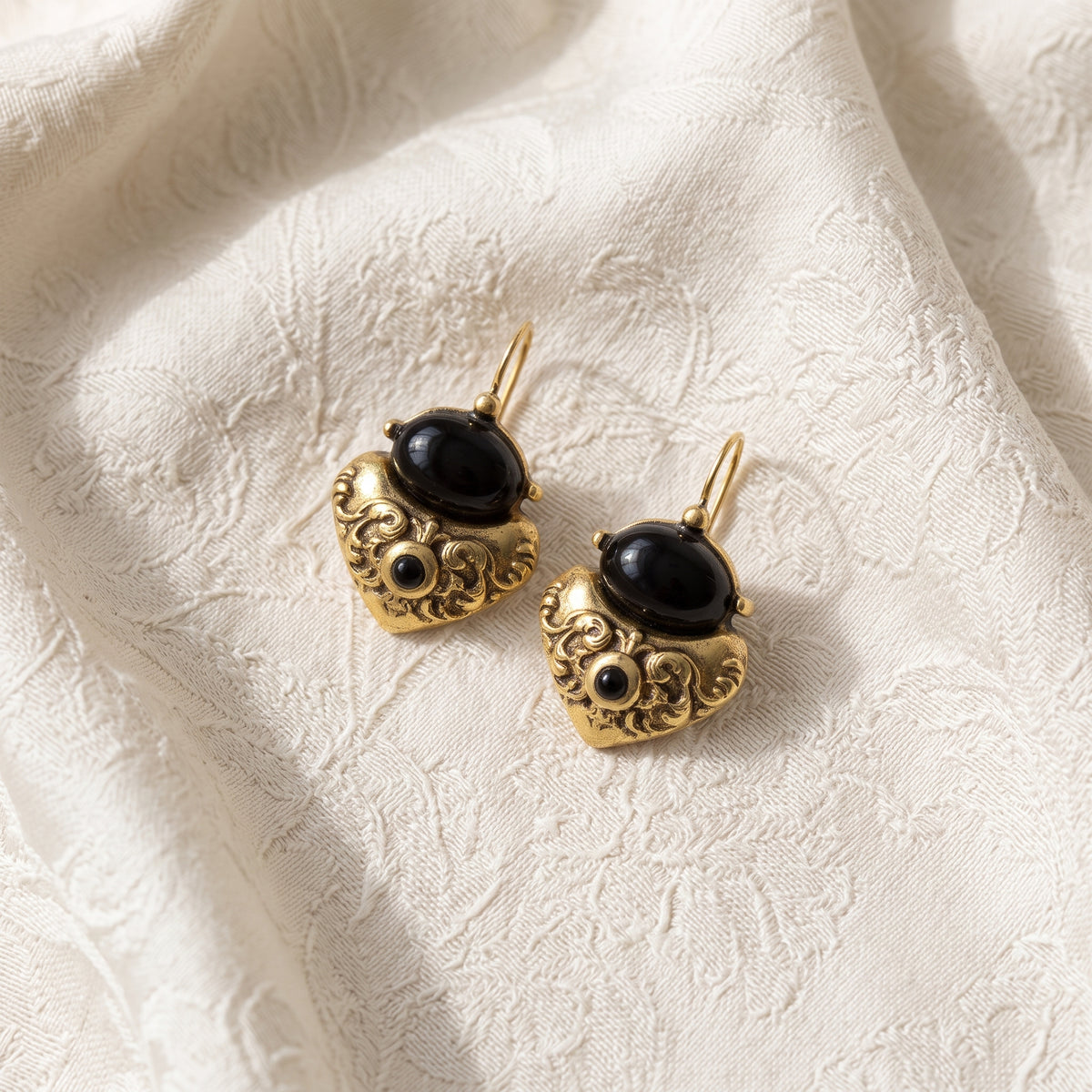 Antiquities Couture Black Corinthian Sculpted Heart Drop Earrings resting on white laced fabric