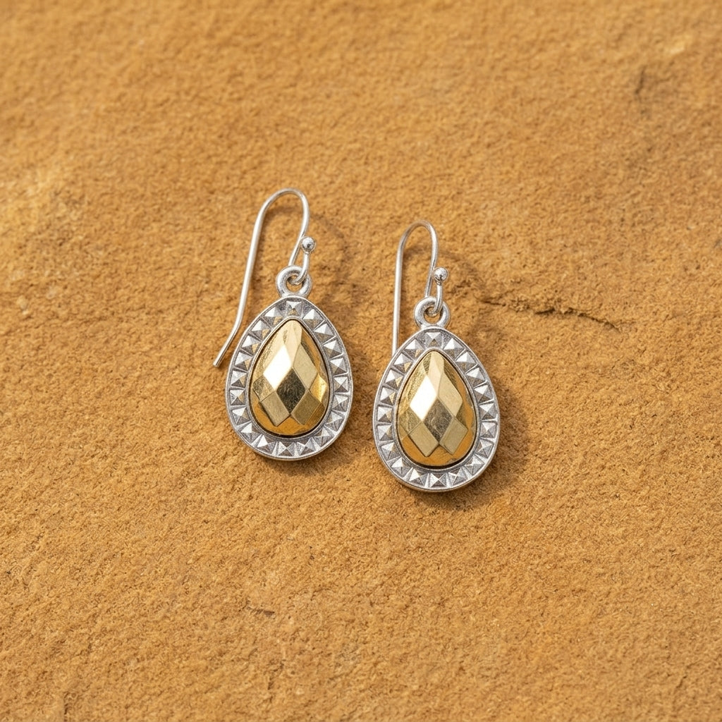 A pair of teardrop-shaped drop earrings with faceted gold tone centers and silver settings, displayed on a textured brown surface.