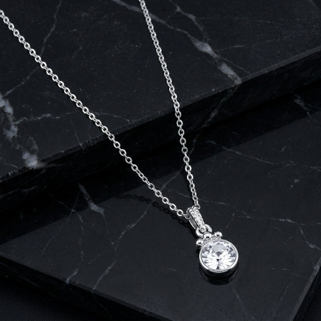 A silver chain necklace with a round clear crystal pendant, displayed on a black marble surface.