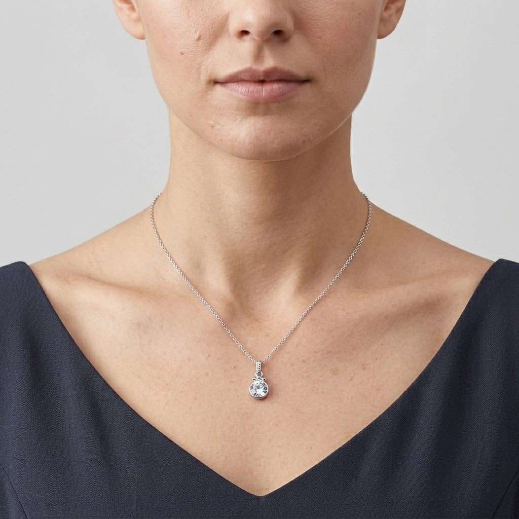 A close-up of a model wearing a delicate silver chain necklace with a round shaped clear crystal pendant, styled with a dark navy blue V-neck top against a light gray background.