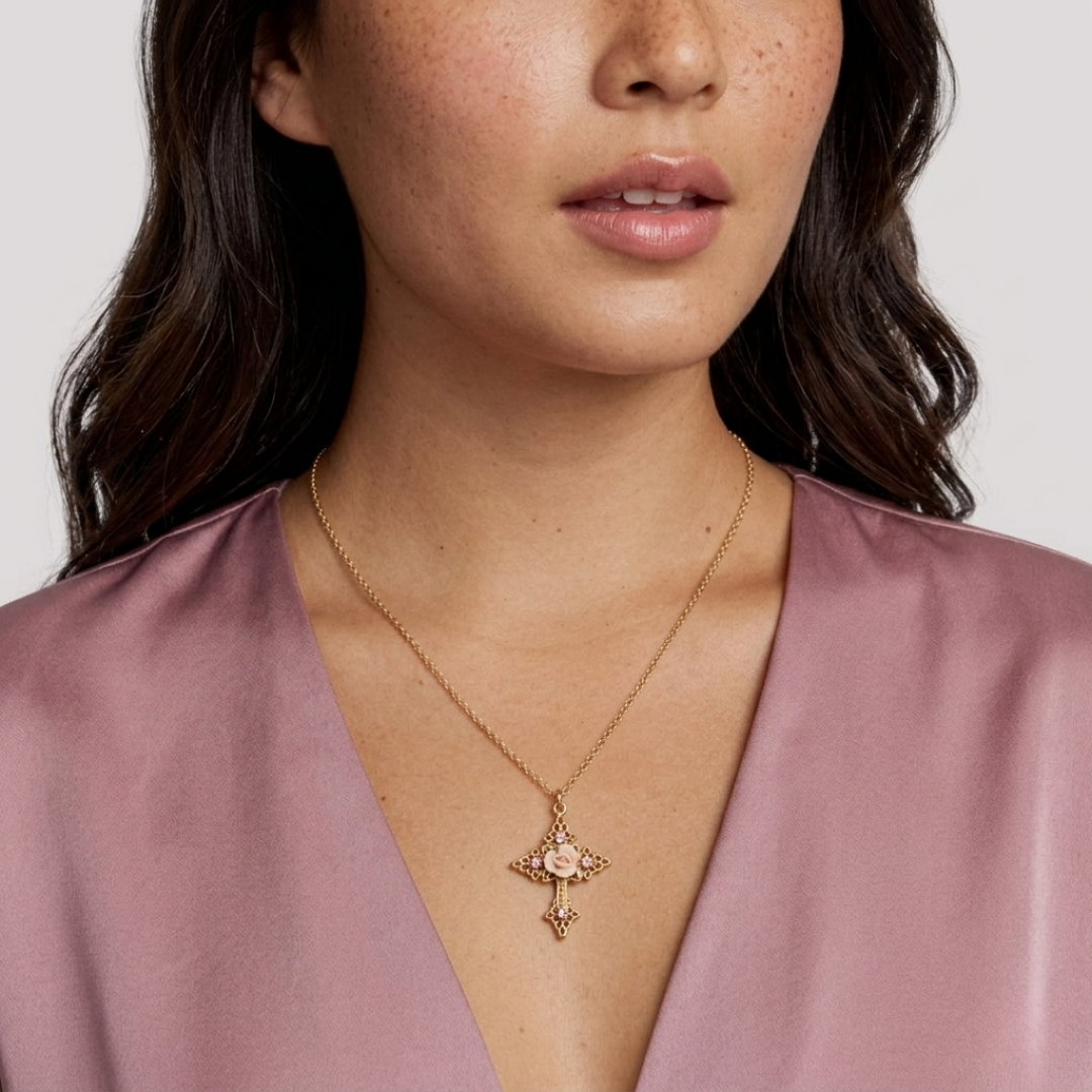 Close-up of a woman wearing a gold cross necklace with faux pearl accents and a pink porcelain rose pendant against a plain background