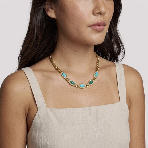 Woman wearing a gold necklace with turquoise and green stones against a plain background