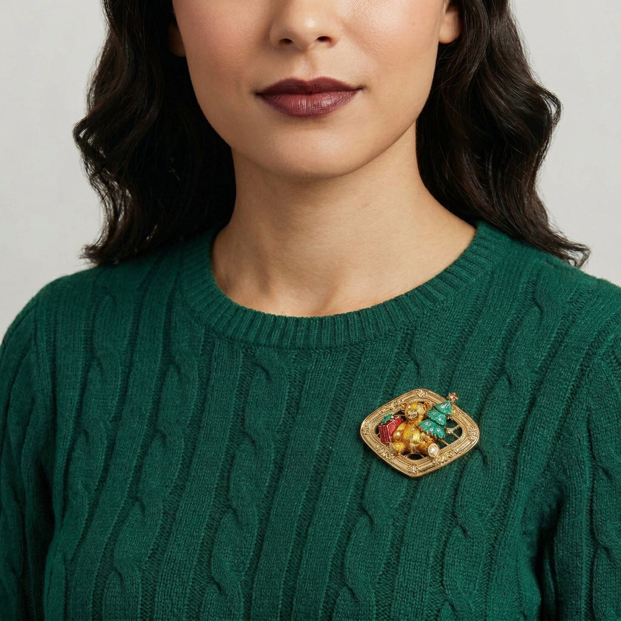 Woman wearing a green sweater with a decorative teddy bear holding a gift next to a Christmas tree brooch on a plain background