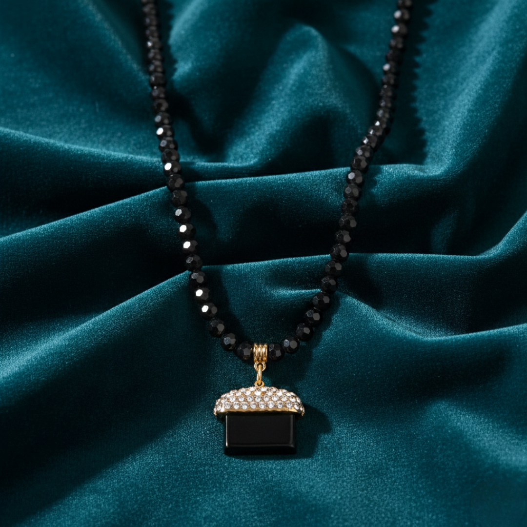 Necklace with a black beaded chain and a black pendant with a gold and crystal accent on a teal fabric background.