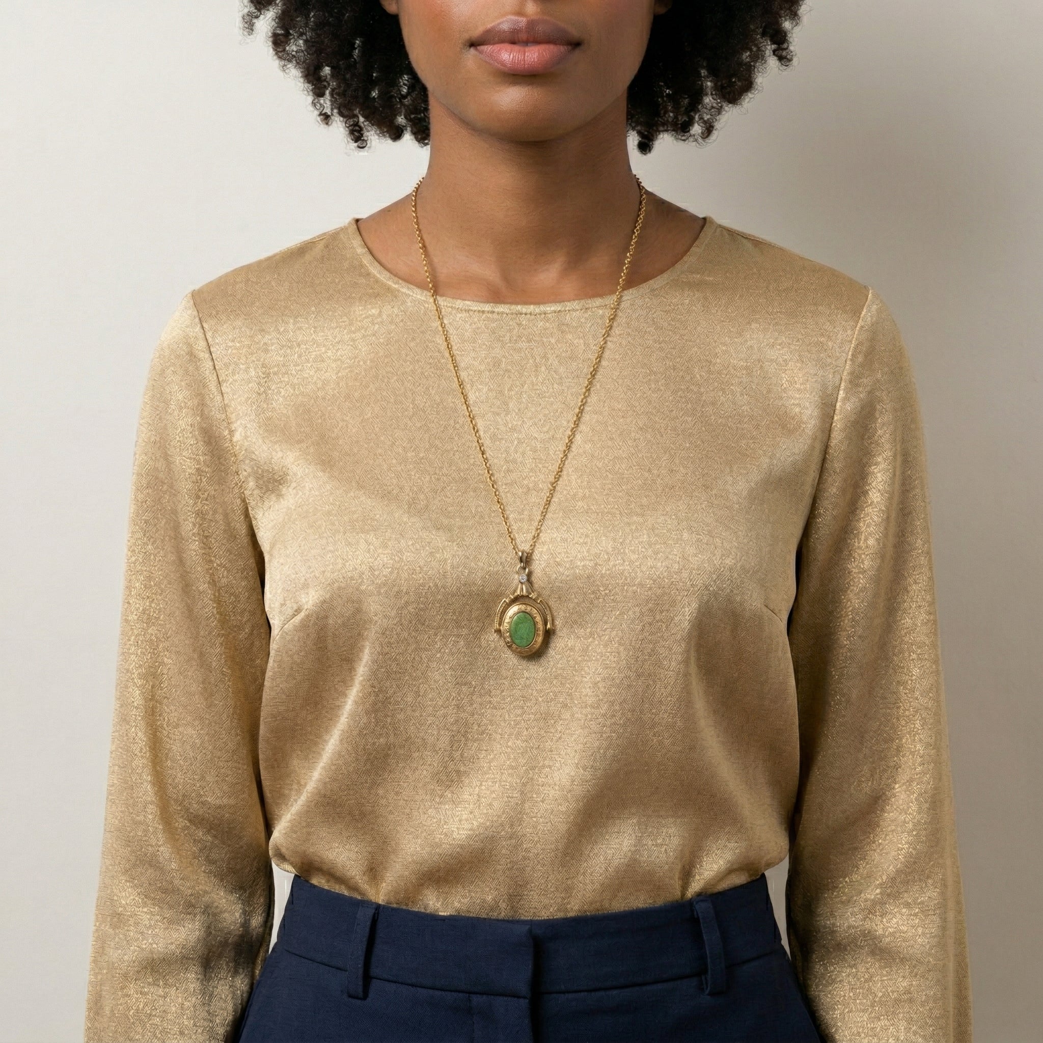 Person wearing a gold long-sleeve top and dark pants with a necklace featuring a green gemstone locket pendant.