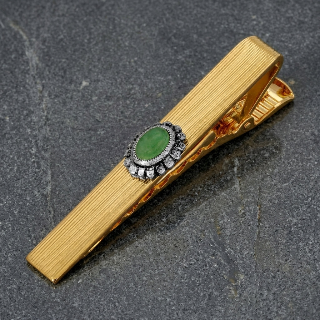 Gold tone men's tie clip with a green semi-precious stone accent resting on smooth stone surface