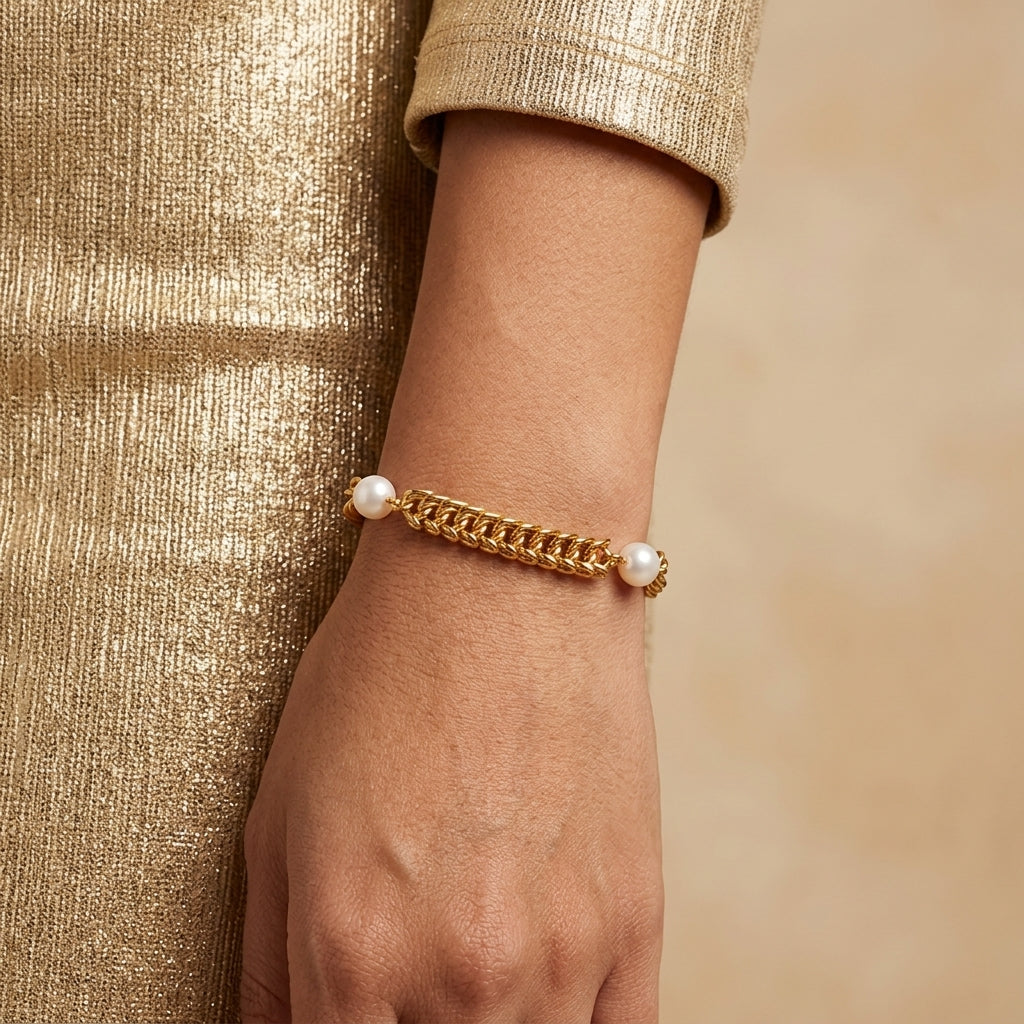 A gold chain bracelet with white pearl accents, shown worn on a person's wrist against a warm beige background.