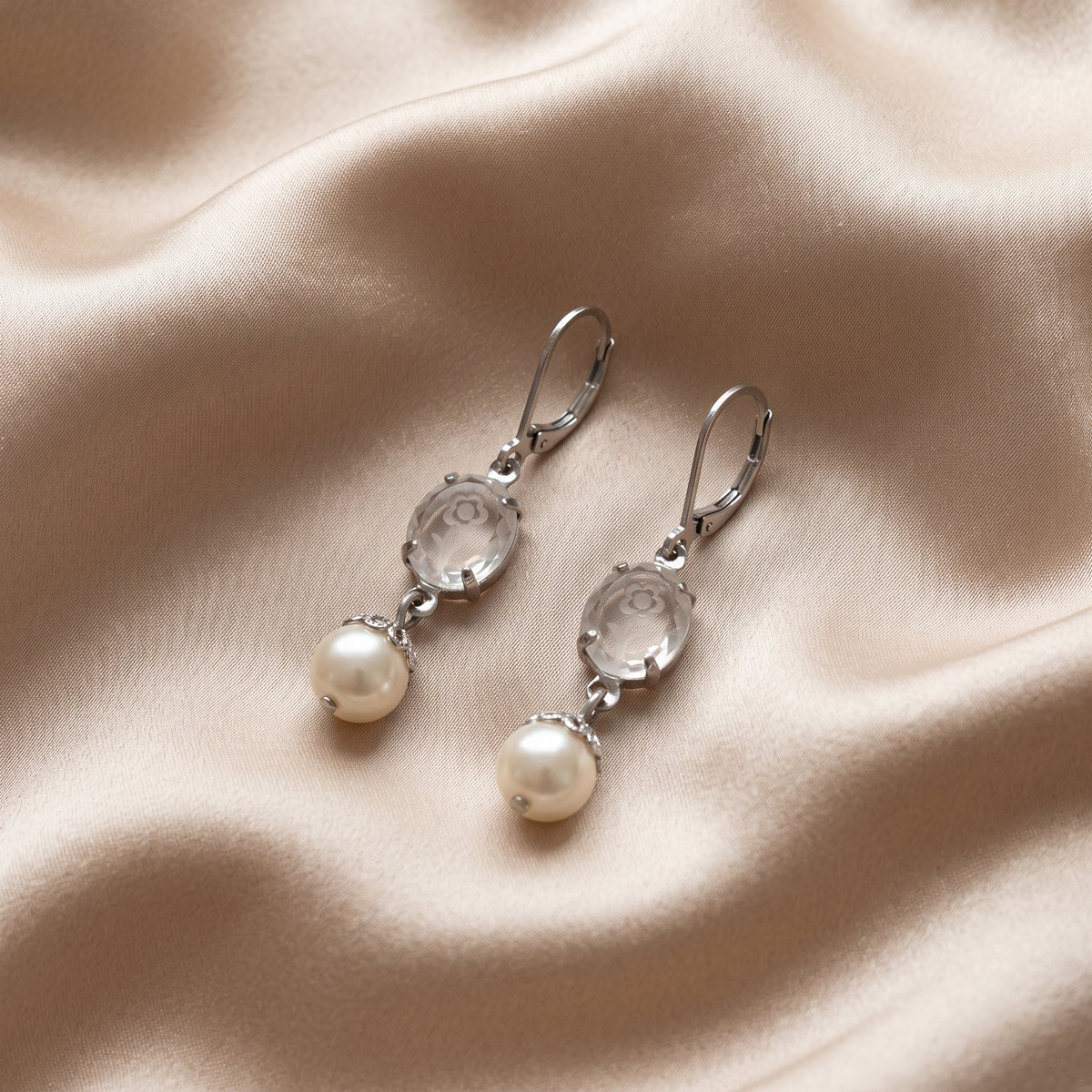 Silver Flower Engraved Crystal & Round Faux Pearl Drop Earrings displayed on a draped beige satin fabric background.