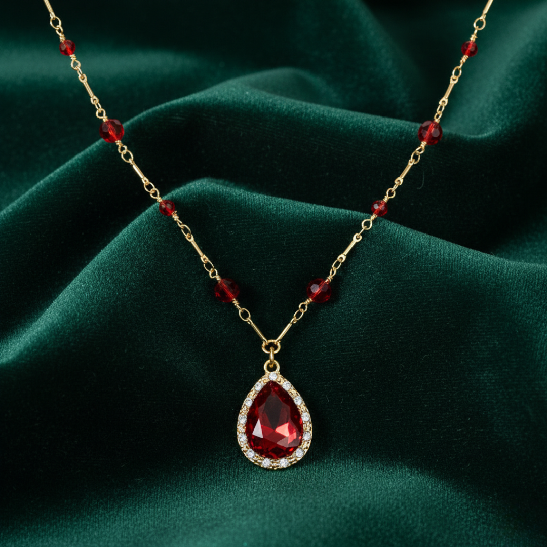 Gold necklace with a red teardrop pendant on a green fabric background.