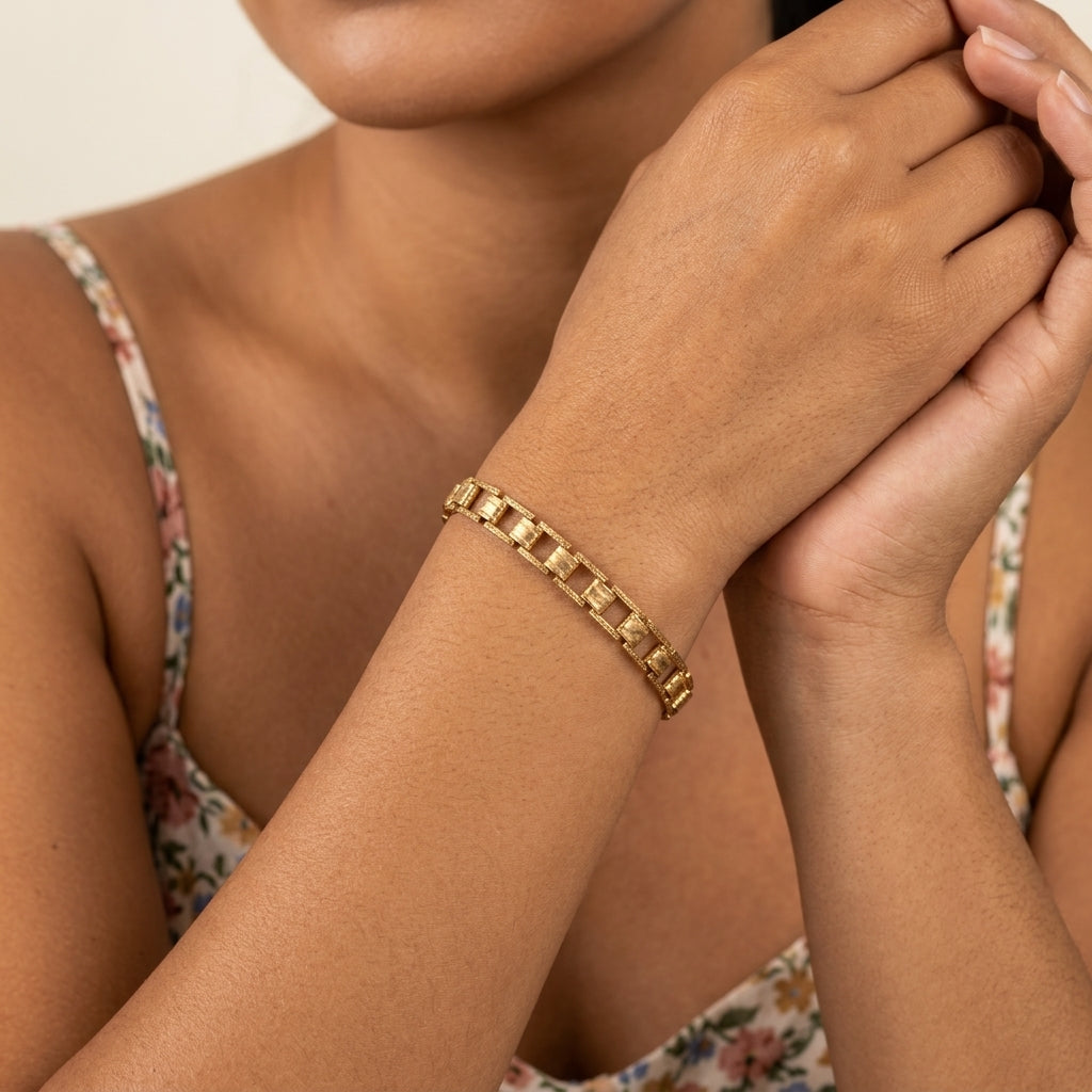 A close-up of a woman's wrist wearing a gold chain link bracelet, with her hands clasped in front. She is wearing a floral-patterned spaghetti-strap top against a clean, light beige background.