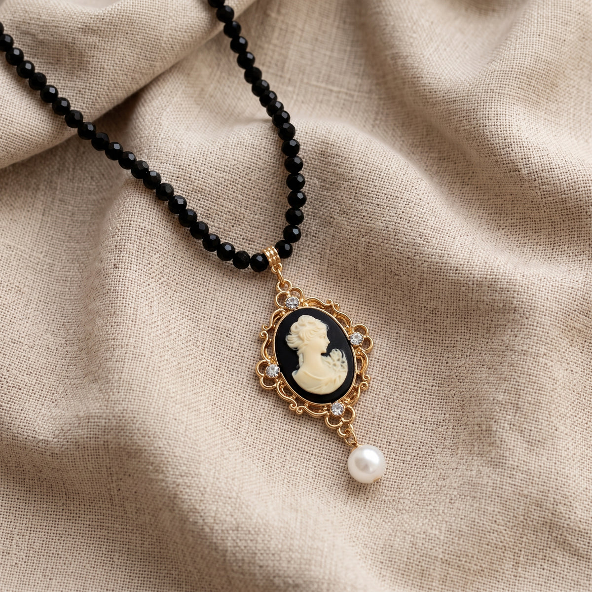 A black beaded necklace with a vintage-style oval pendant featuring an ivory-colored classical cameo portrait, surrounded by a gold-tone frame with small clear stones and a dangling faux pearl, displayed on draped beige linen fabric.