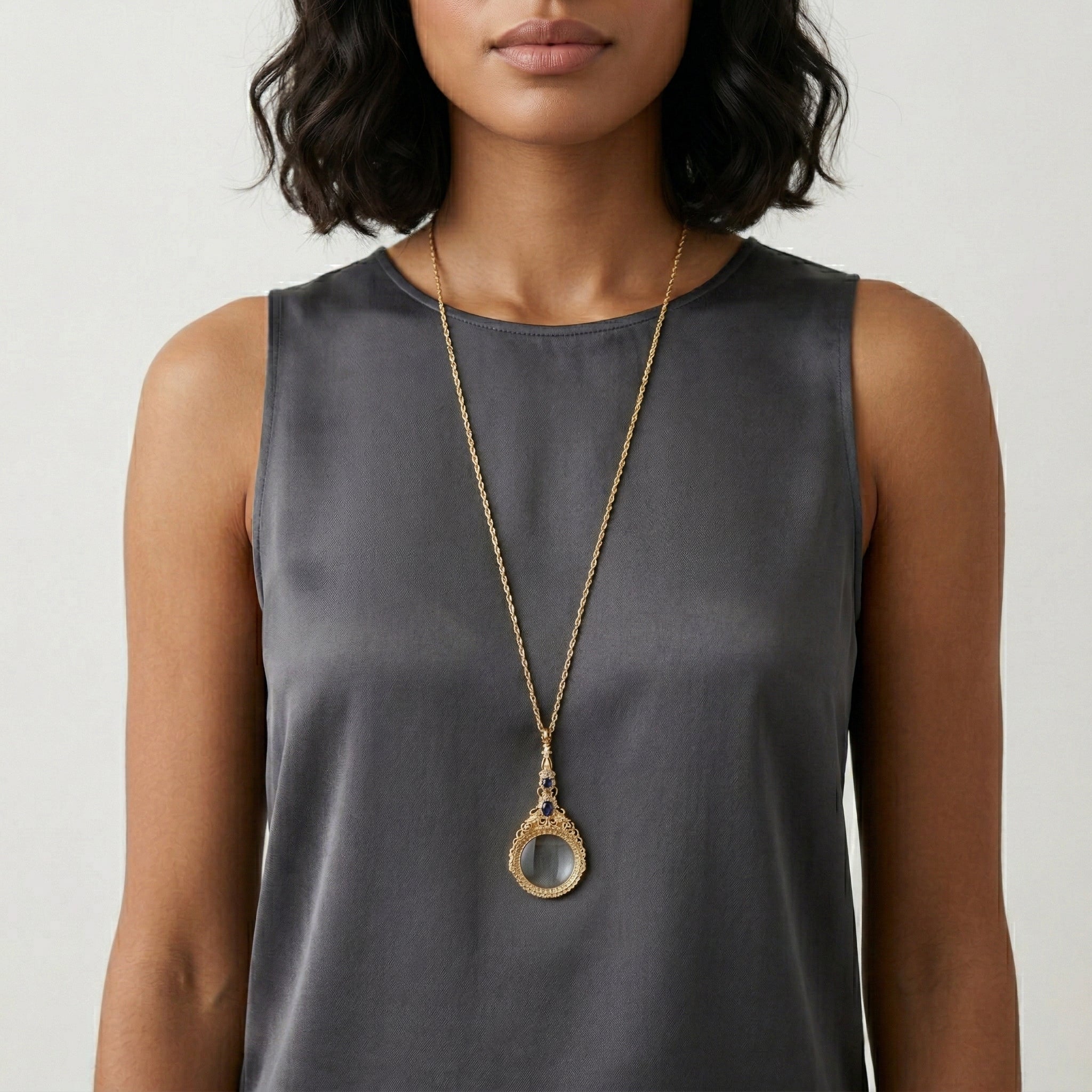 Woman wearing a long gold necklace with a magnifying glass pendant accented with blue crystals on a plain background