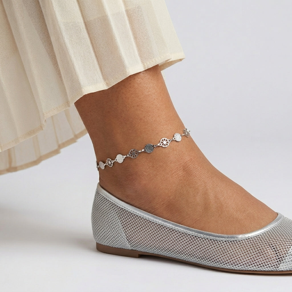 Silver anklet on a foot wearing a silver flat shoe with a plain background