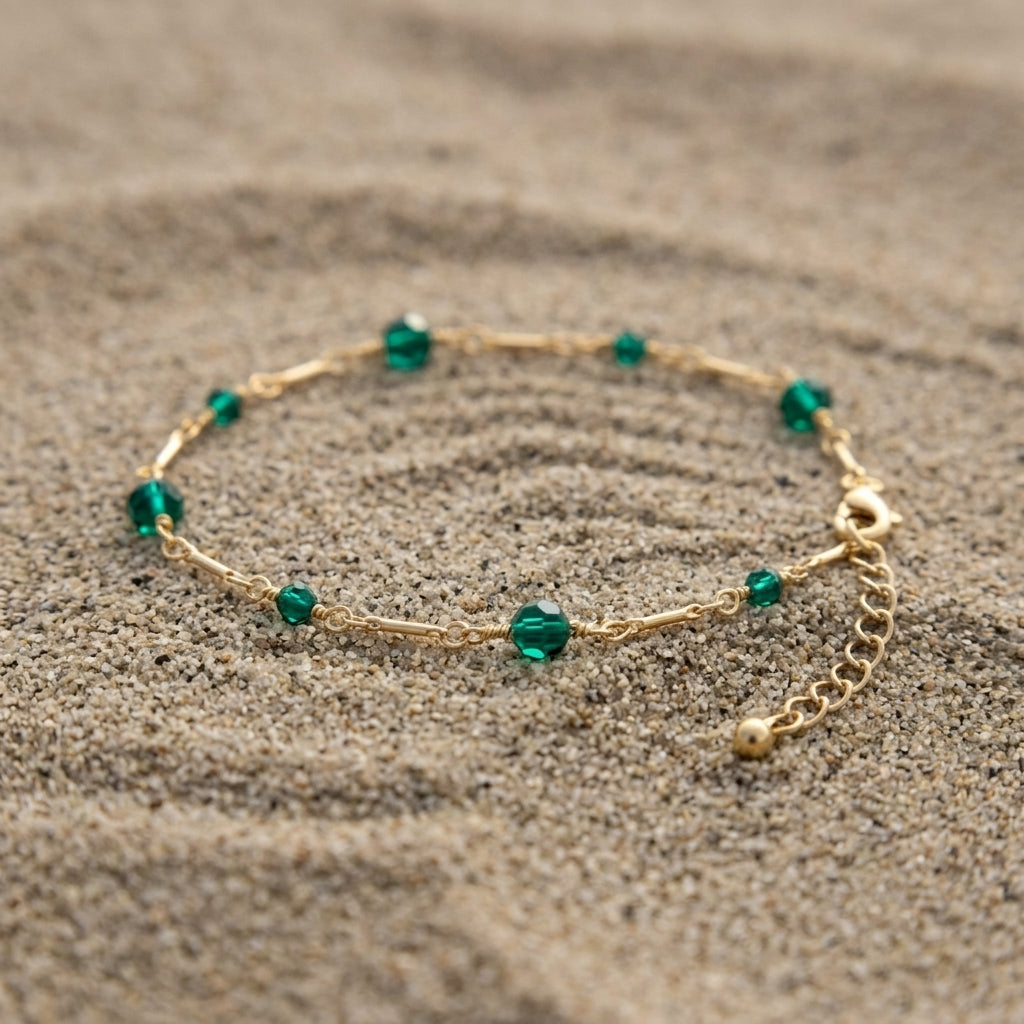 A delicate gold chain anklet with small faceted green crystal beads, displayed on a textured sandy surface.
