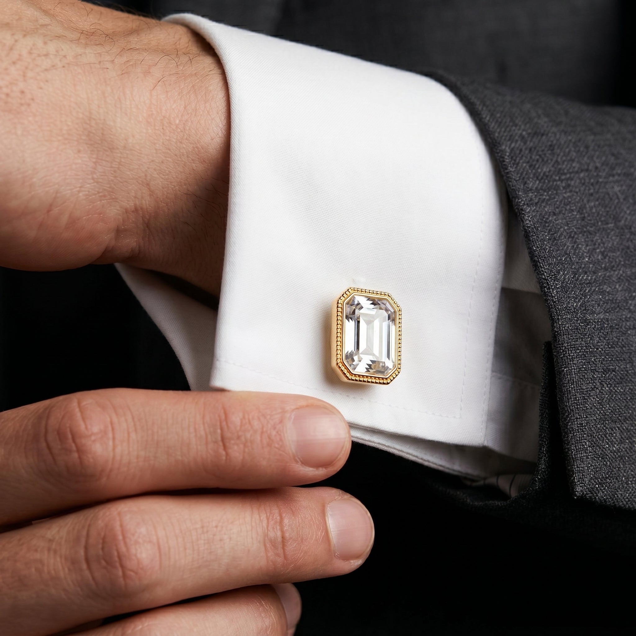 Close-up of a cufflink with a clear crystal and gold tone frame on a white cuff against a dark suit background