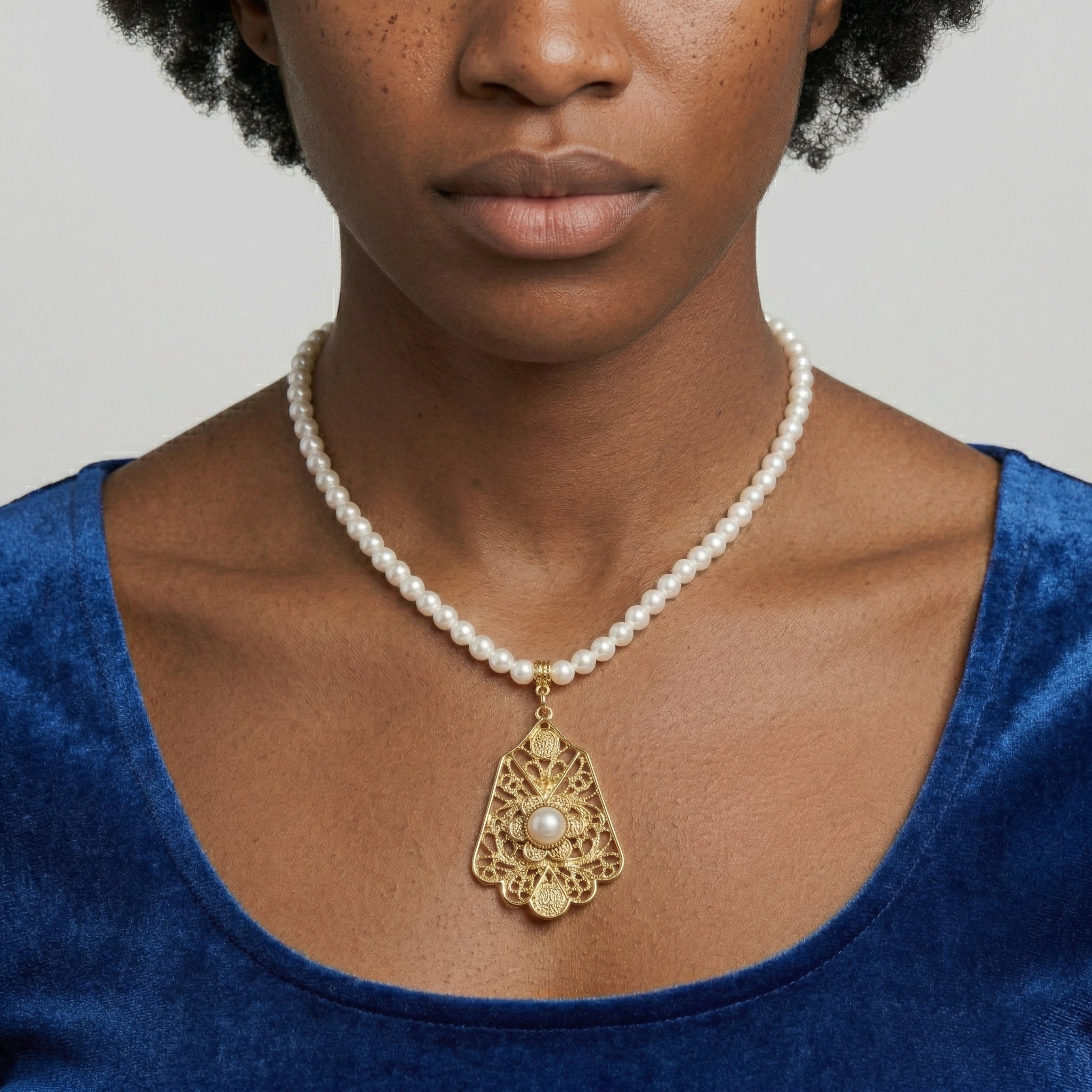 Pearl necklace with a gold pendant worn by a person in a blue top against a neutral background