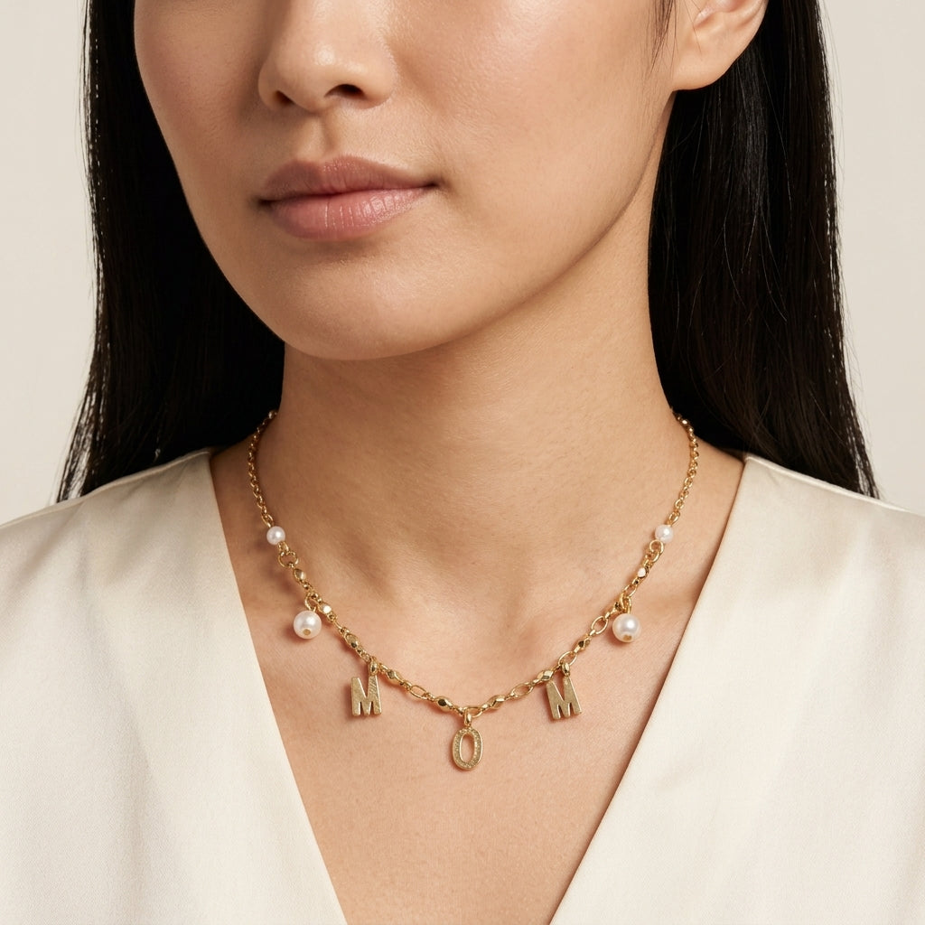Close-up of a woman wearing a gold tone "MOM" initialed necklace with white faux pearl accents on a neutral background