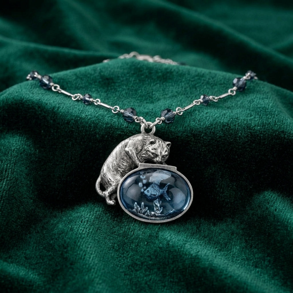 Silver tone Cat And Blue Fish Pendant Necklace with blue bead accents resting on a dark green fabric surface