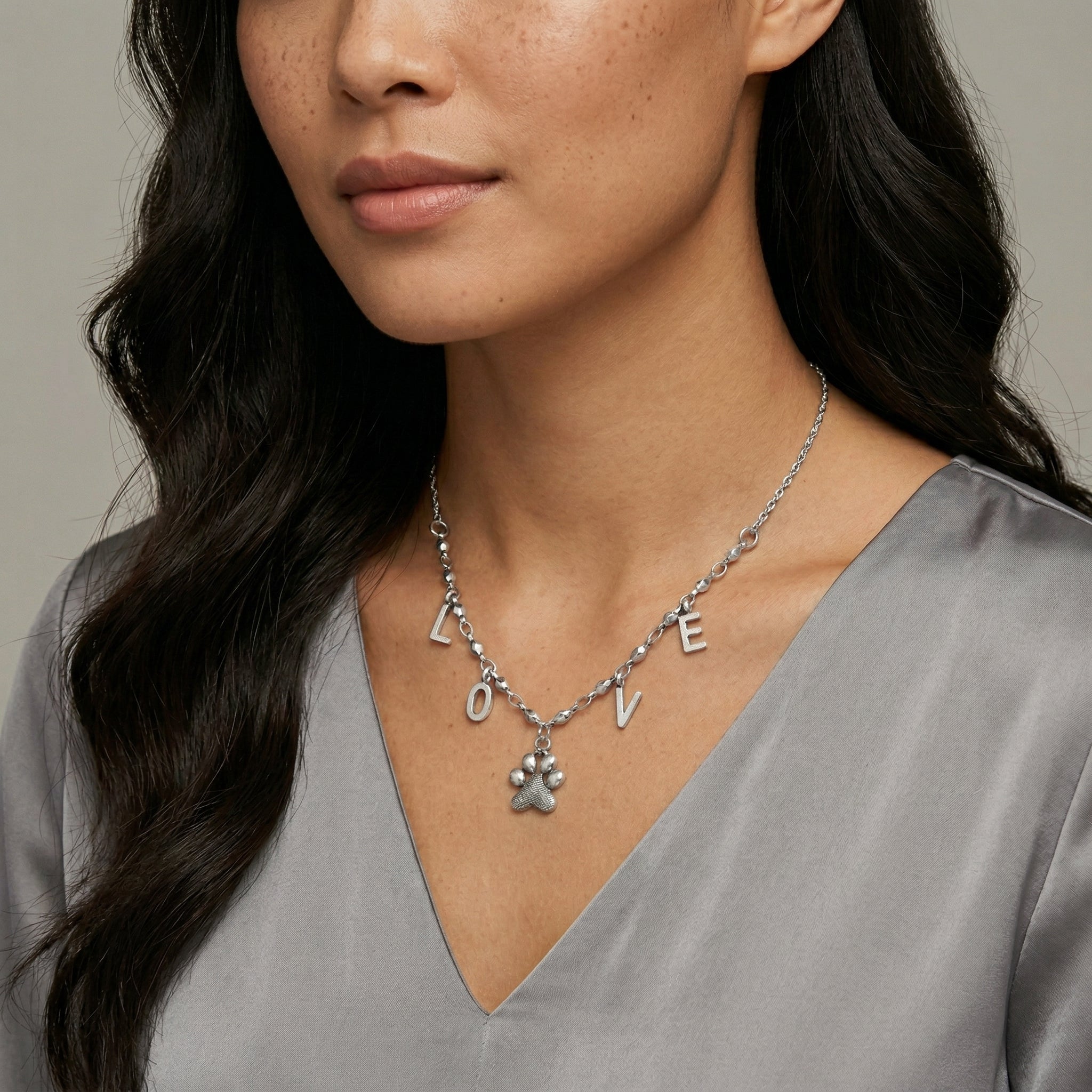 Woman wearing a silver necklace with 'LOVE' and paw print charms on a neutral background