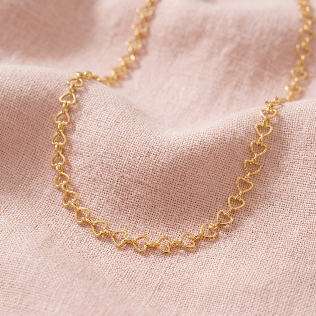 A delicate gold chain necklace featuring repeating interlocking arrow links, draped on a soft pink textured fabric background.