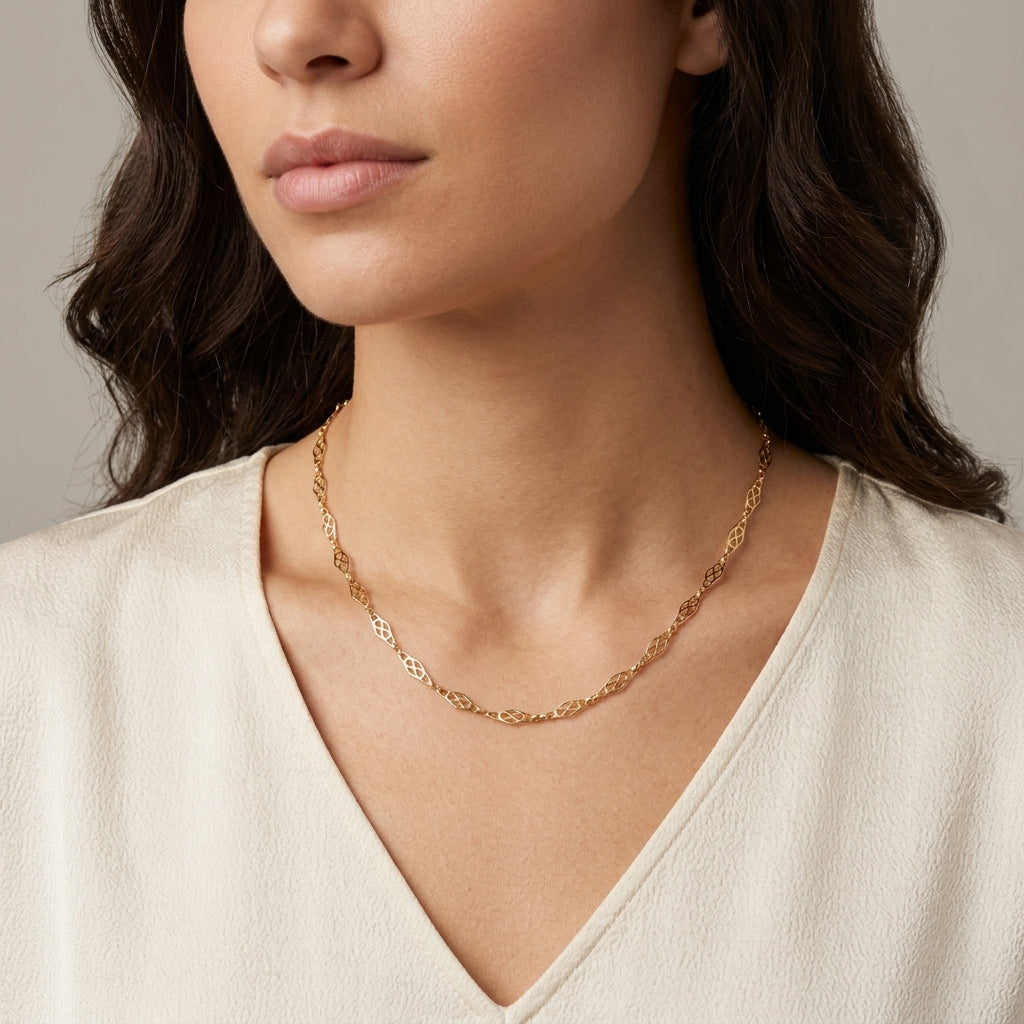 A close-up of a woman wearing a delicate gold chain necklace with a textured, openwork design. She is wearing a cream-colored V-neck top against a neutral gray background.