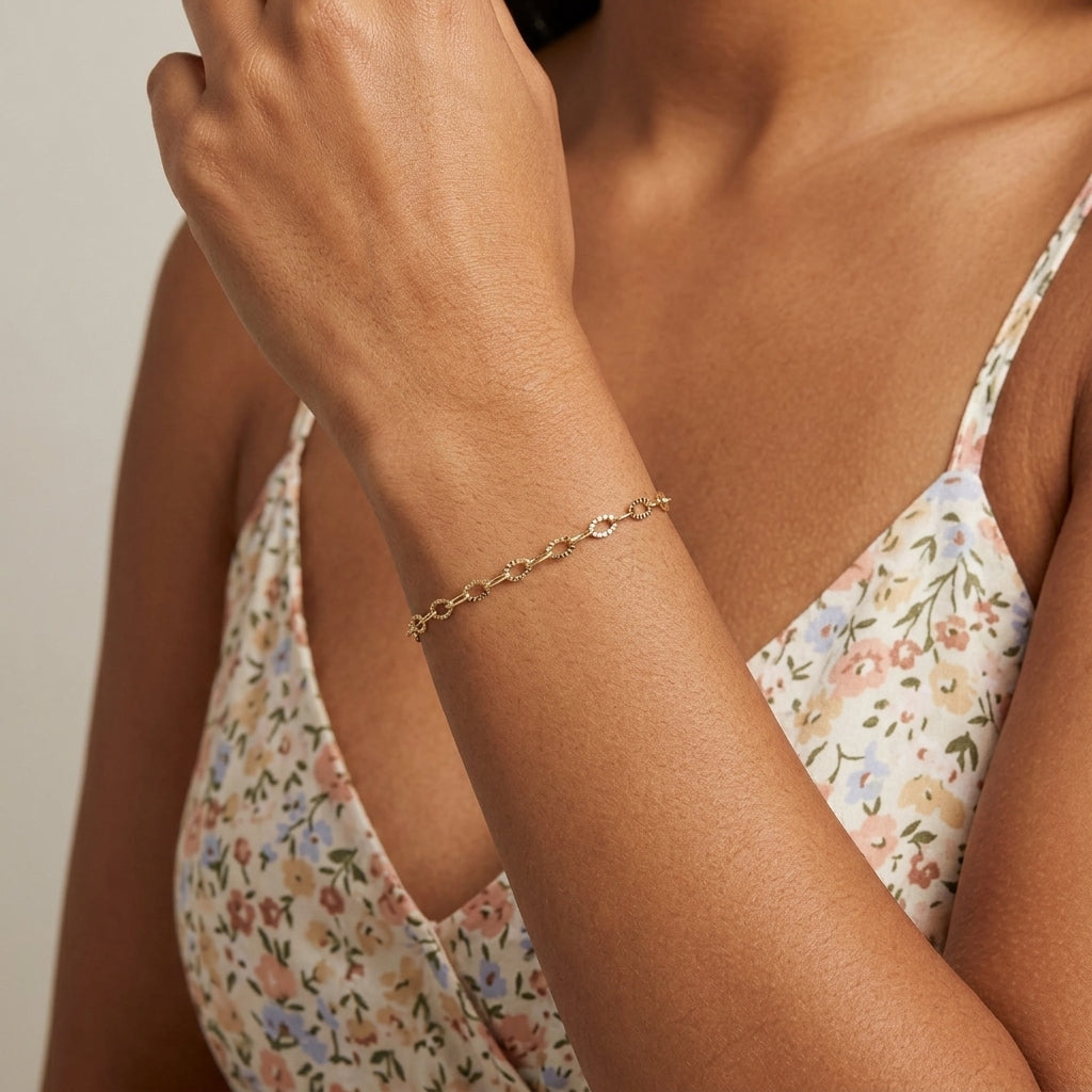 A delicate gold chain bracelet worn on a woman's wrist, shown in a close-up lifestyle shot. The model is wearing a floral-patterned camisole against a neutral light gray background.