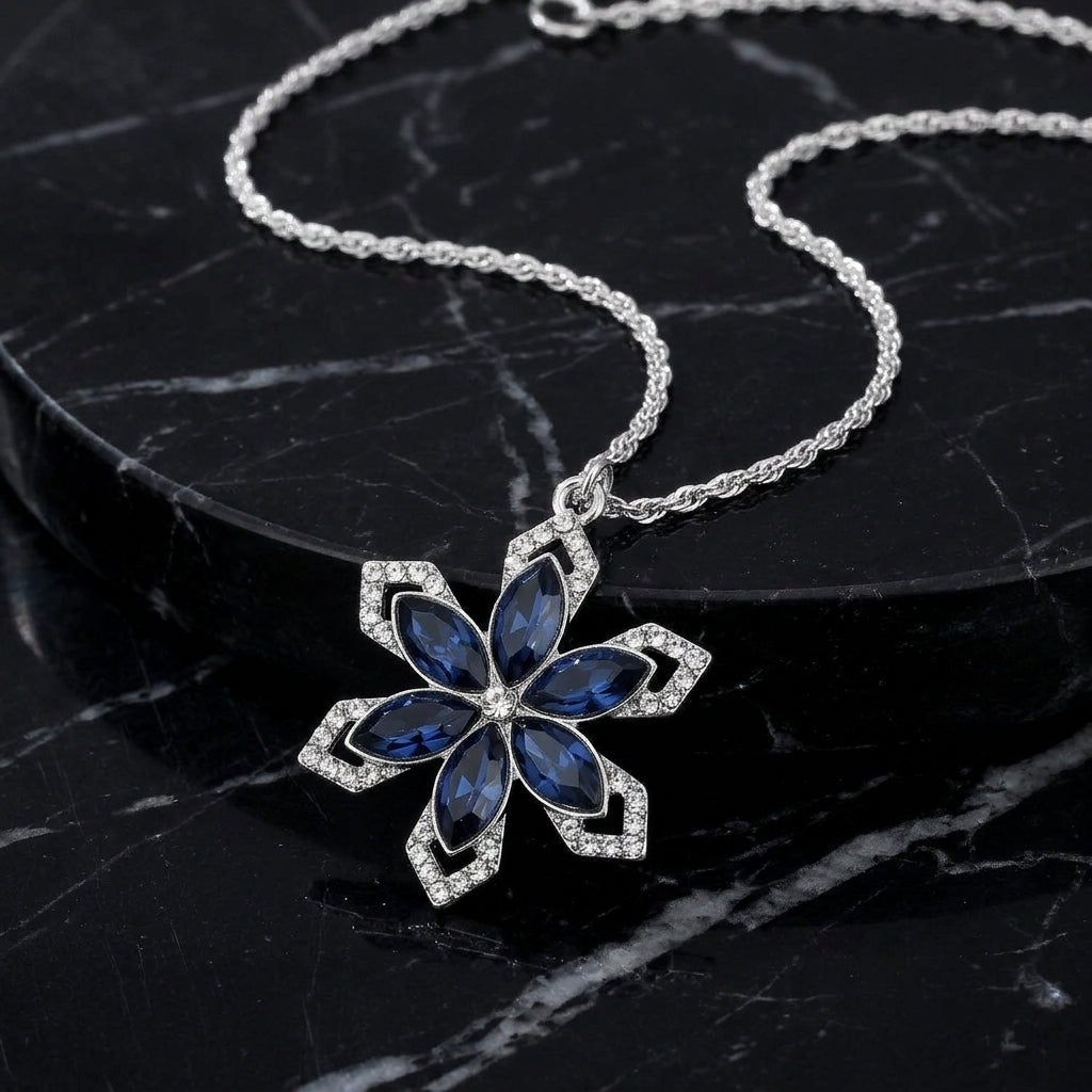 Sapphire blue crystal snowflake pendant necklace that has a silver tone chain resting on a black marble stone surface