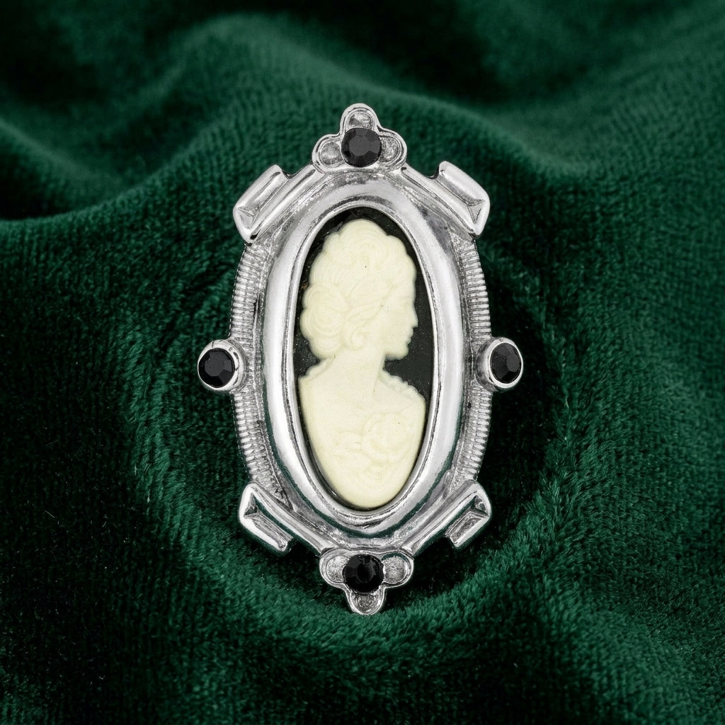 Black cameo brooch with black crystal accents and a silver tone frame resting on dark green fabric
