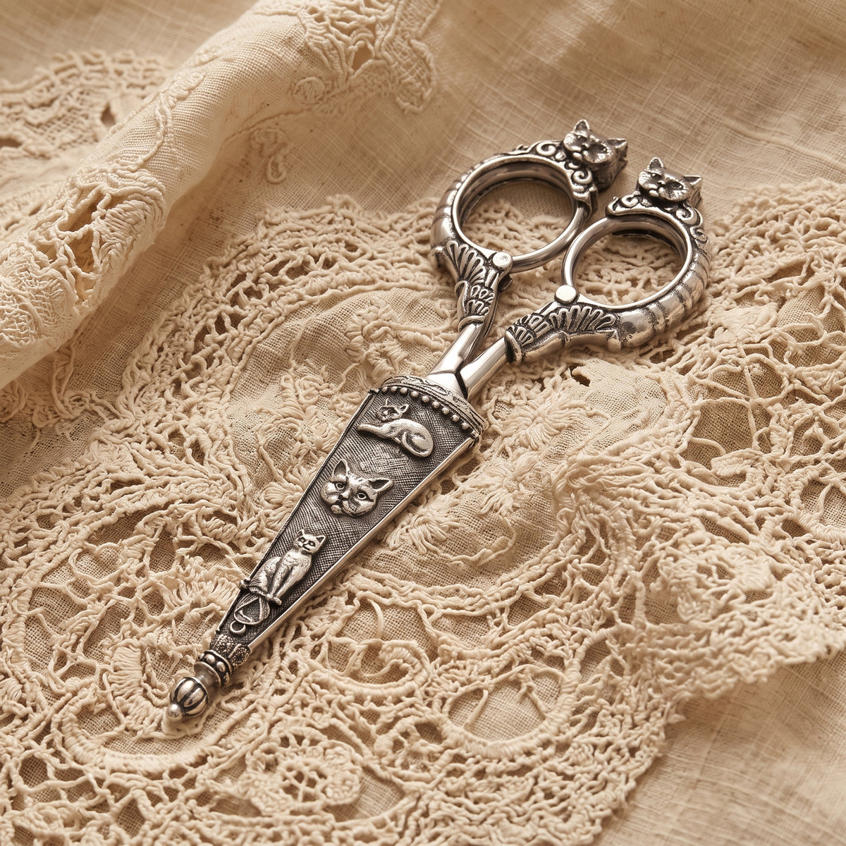 Decorative silver scissors with cat-shaped handles and embossed cat motifs on the blade, placed on beige lace fabric.