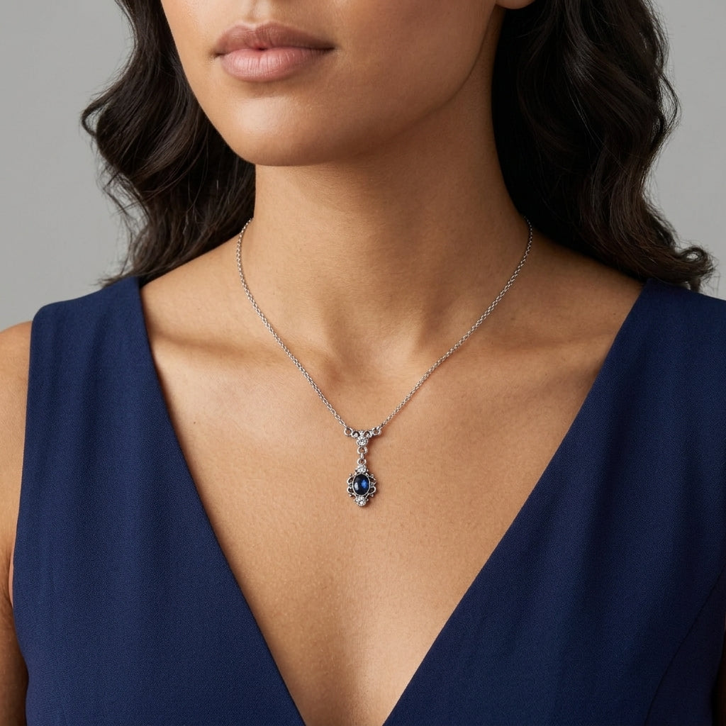 A close-up of a woman wearing a delicate silver pendant necklace with a dark blue oval gemstone and small clear accent stones, paired with a navy blue dress.