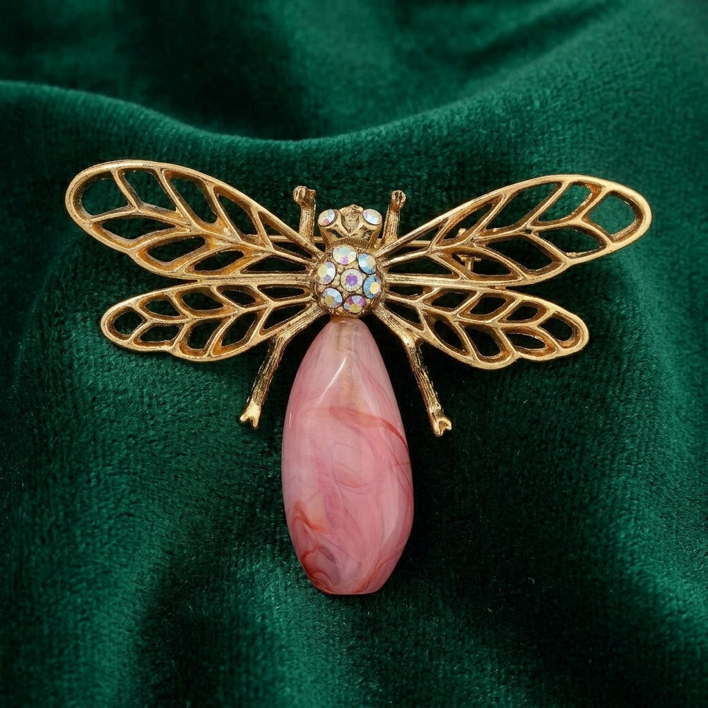 Gold tone dragonfly-shaped brooch with a pink stone and AB crystals on a green fabric background