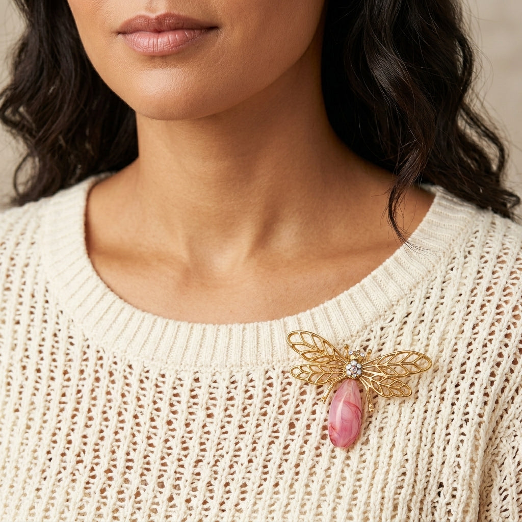 Close-up of a person wearing a beige sweater with a decorative pink dragonfly brooch featuring a pink stone and gold tone design.
