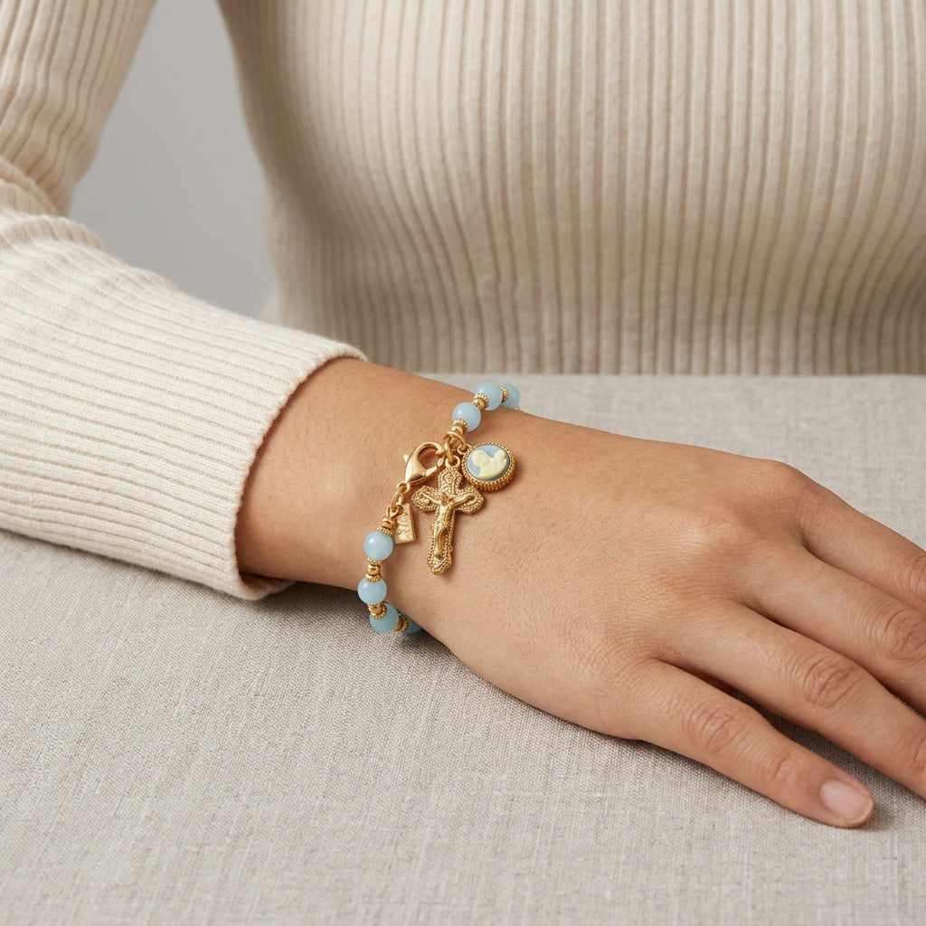 Hand wearing a gold bracelet with blue beads and a crucifix charm on a neutral background
