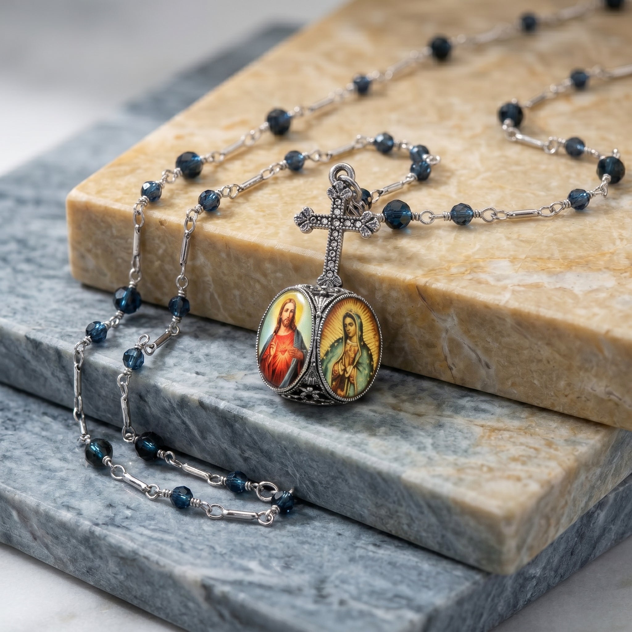 Silver tone cross rosary-style necklace with Montana Blue beads on the chain and a pendant with Jesus and the Virgin of Guadalupe motifs resting on a stone surface.