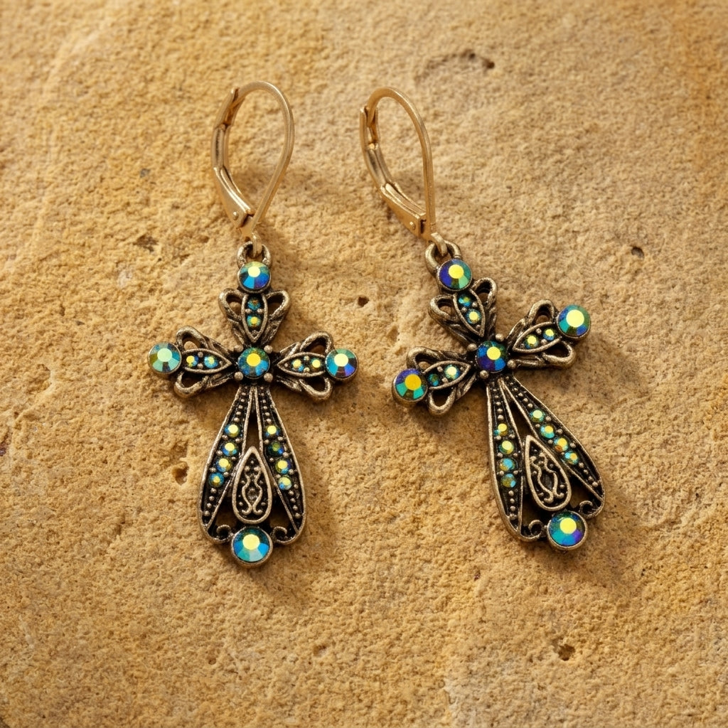 A pair of ornate filigree cross drop earrings with iridescent blue-green crystal stones on a textured tan stone background.