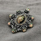 An ornate vintage-style brooch featuring a central oval jet black stone surrounded by filigree, iridescent green and iridescent pink stones, and small AB crystals displayed on a textured gray fabric background.