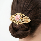Gold hair barrette with a pink porcelain rose flower on a bun against a white background