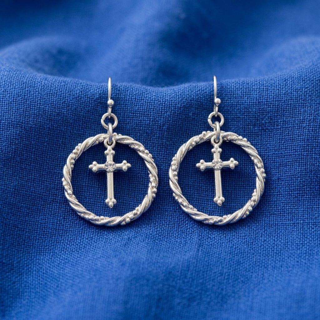 A pair of silver-toned drop earrings featuring a small cross inside a twisted rope-style circular frame, displayed on a textured blue fabric background.