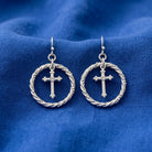 A pair of silver-toned drop earrings featuring a small cross inside a twisted rope-style circular frame, displayed on a textured blue fabric background.