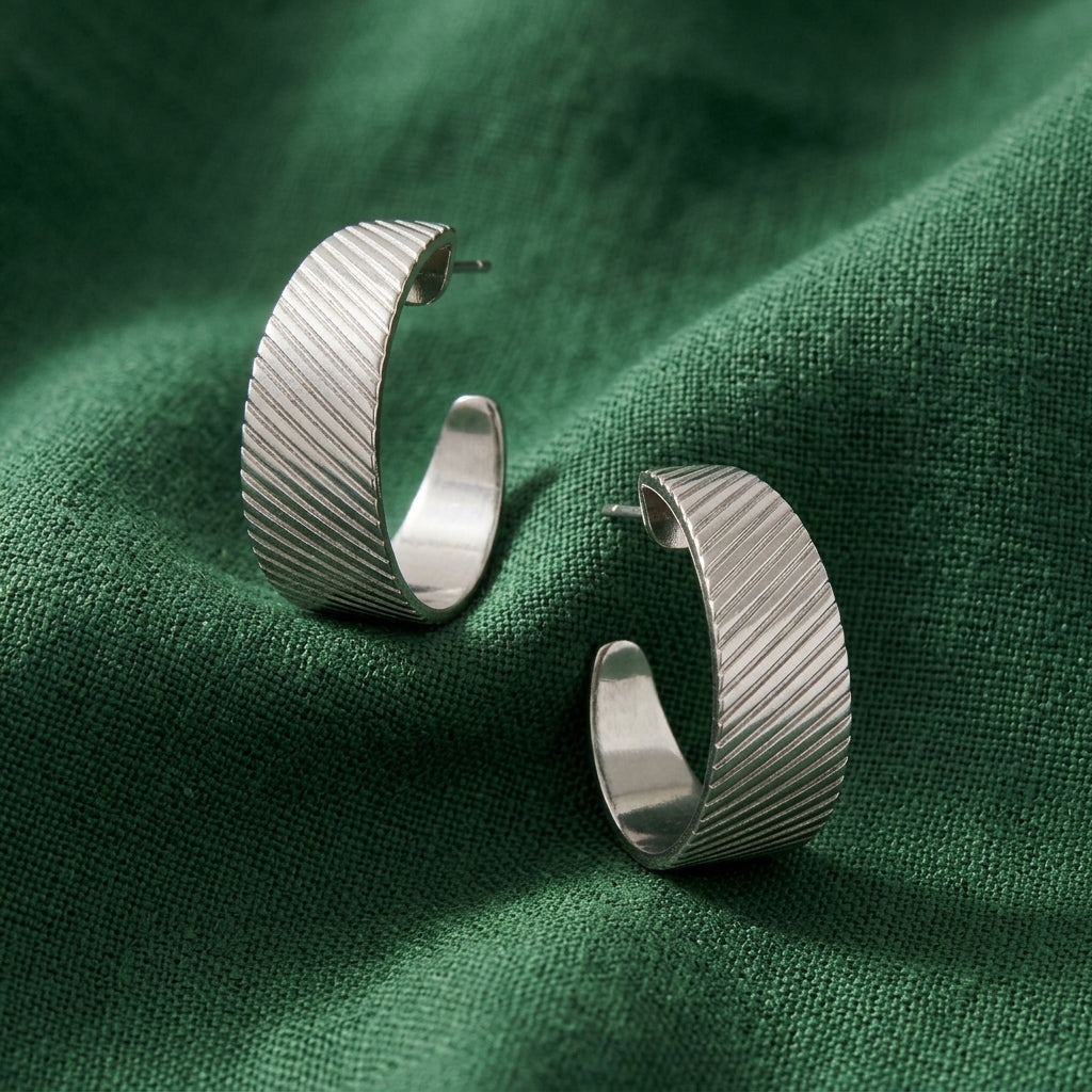 A pair of silver hoop earrings with a textured, ribbed diagonal pattern, displayed on a draped dark green fabric background.