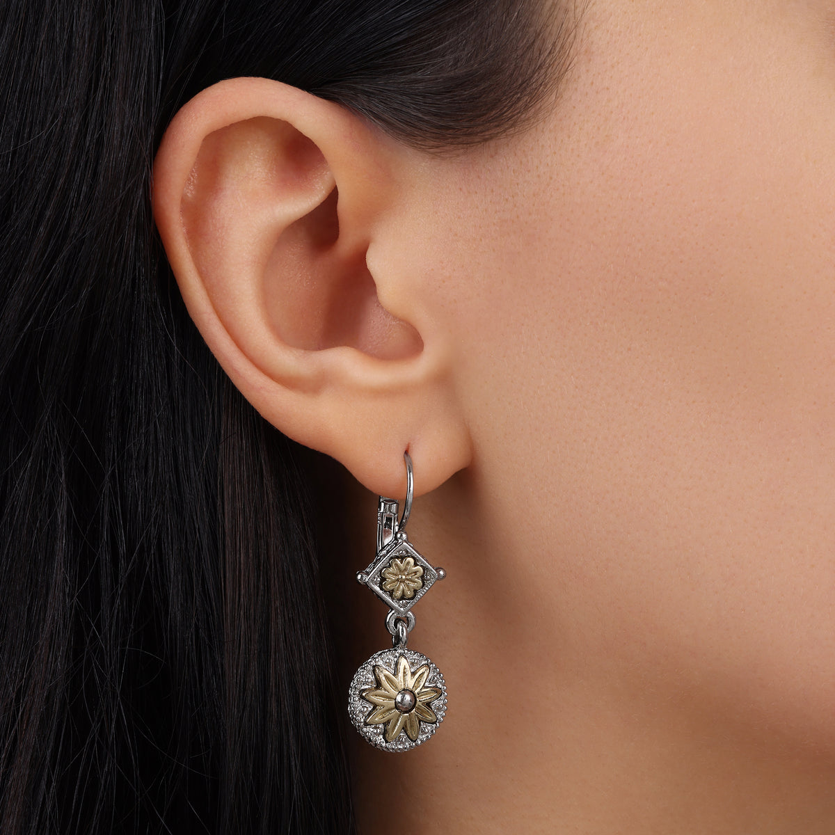 A close-up of a model's ear wearing a silver tone dangle earring with a diamond-shaped link, a small flower motif, and a larger circular flower drop.
