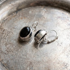 Antiquities Couture Illusions Onyx And Silver Oval Drop Leverback Earrings resting on an antique silver metal dish