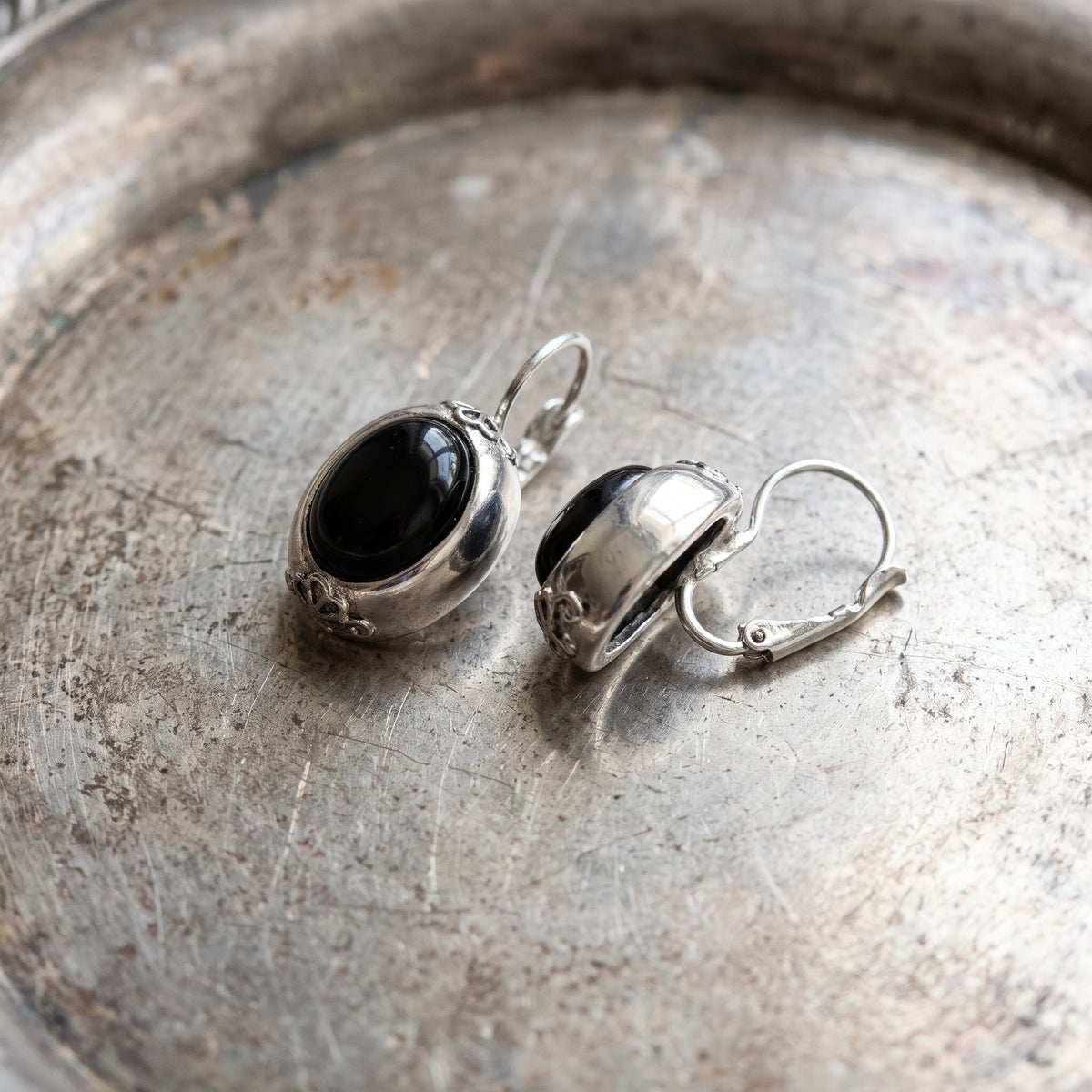 Antiquities Couture Illusions Onyx And Silver Oval Drop Leverback Earrings resting on an antique silver metal dish