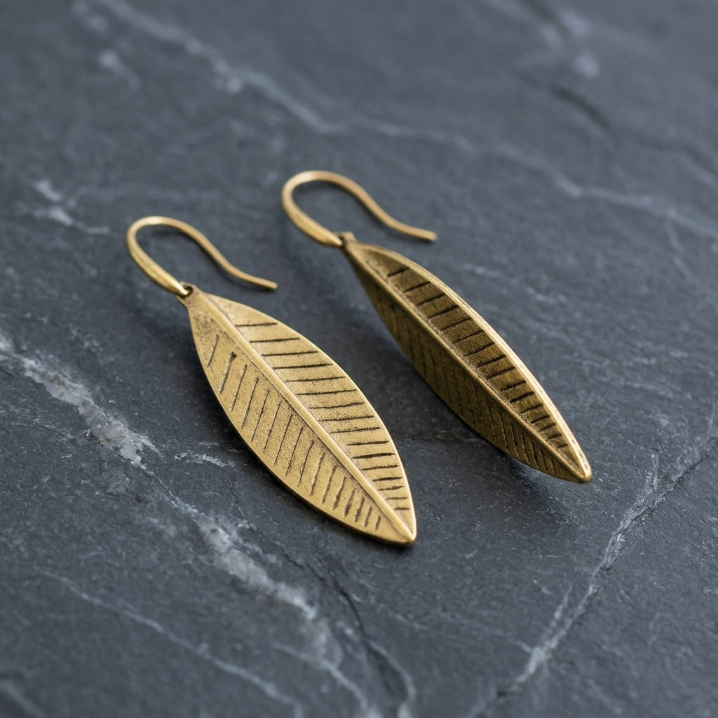 A pair of gold-toned drop earrings shaped like elongated leaves with engraved vein details, resting on a dark gray slate surface.