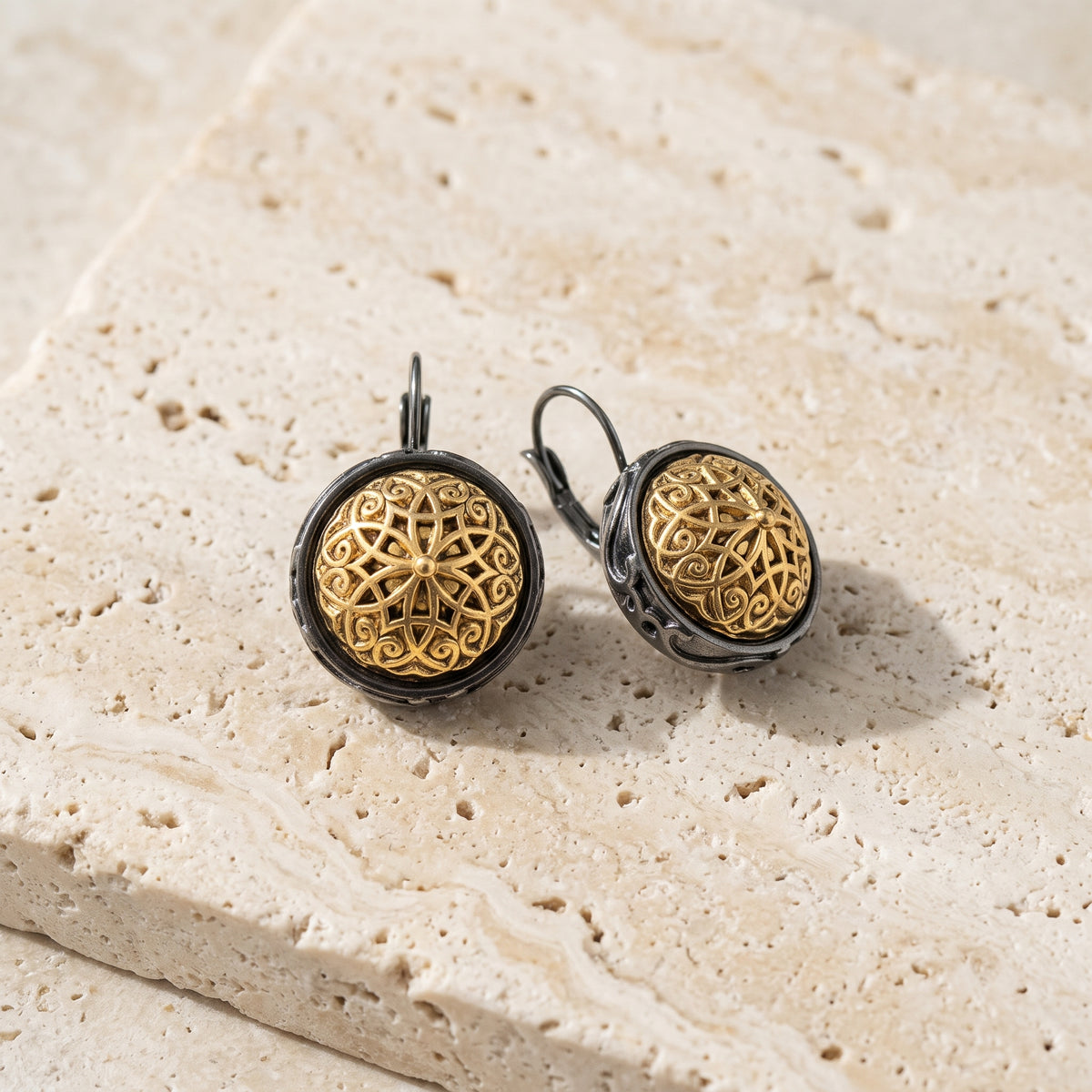 Antiquities Couture Patras Jet Black Gold Button Leverback Earrings displayed on a textured beige stone surface.