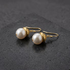 White faux pearl earrings with a gold tone fish hook backing resting on a dark stone surface